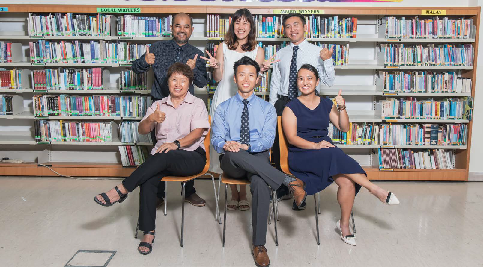 Six formally dressed P.E Teachers pose around a small round table in a library with bookshelves in the background.